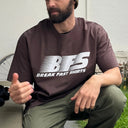 A man wearing a dark brown t-shirt with a bold white 'BFS BREAK FAST SHIRTS' logo on the front, paired with olive green pants, sitting outdoors.