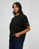 All About Eve Basic Monogram Tee in black cotton, relaxed fit with sleeve logo detail against grey background