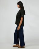 Side view of All About Eve Basic Monogram Tee in black, styled with wide-leg jeans and sandals against grey background