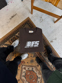 A dark brown crewneck t-shirt with a white 'BTS BREAK FAST SHIRTS' logo is laid flat on a patterned rug, viewed from above with the person's legs and shoes visible around it.