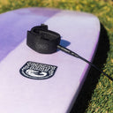 Close-up of a white surfboard with a black leash and 'Creative Army' logo on a grassy background. O&E SOFTBOARD