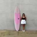 Woman standing next to a pink surfboard against a gray wall. O&E SOFT BOARD