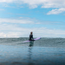 Person in a wetsuit sitting on a purple surfboard in the ocean with a blue sky.